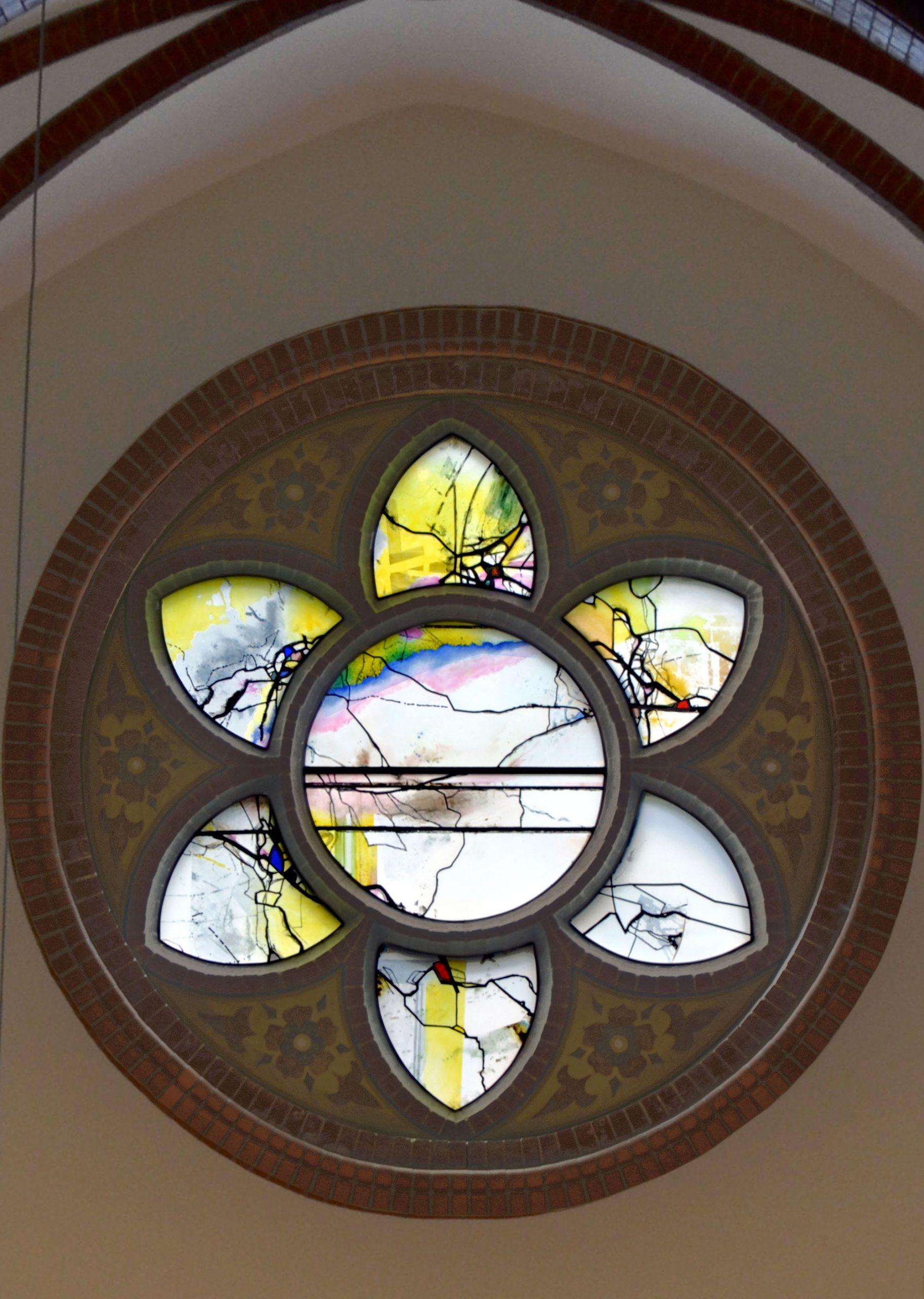 Buntglas-Rosette in einer Kirche mit geometrischen Mustern und natürlichen Motiven in gelblichen und bläulichen Tönen.