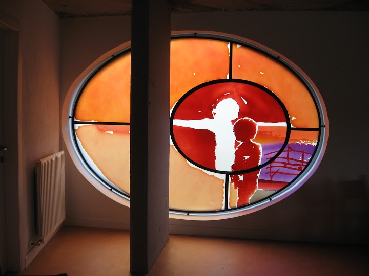 054 – Derix Glasstudios Rundes Kirchenfenster mit silbernem Rahmen und rotem Glas, zentrales Motiv zeigt helle Silhouette eines Menschen.