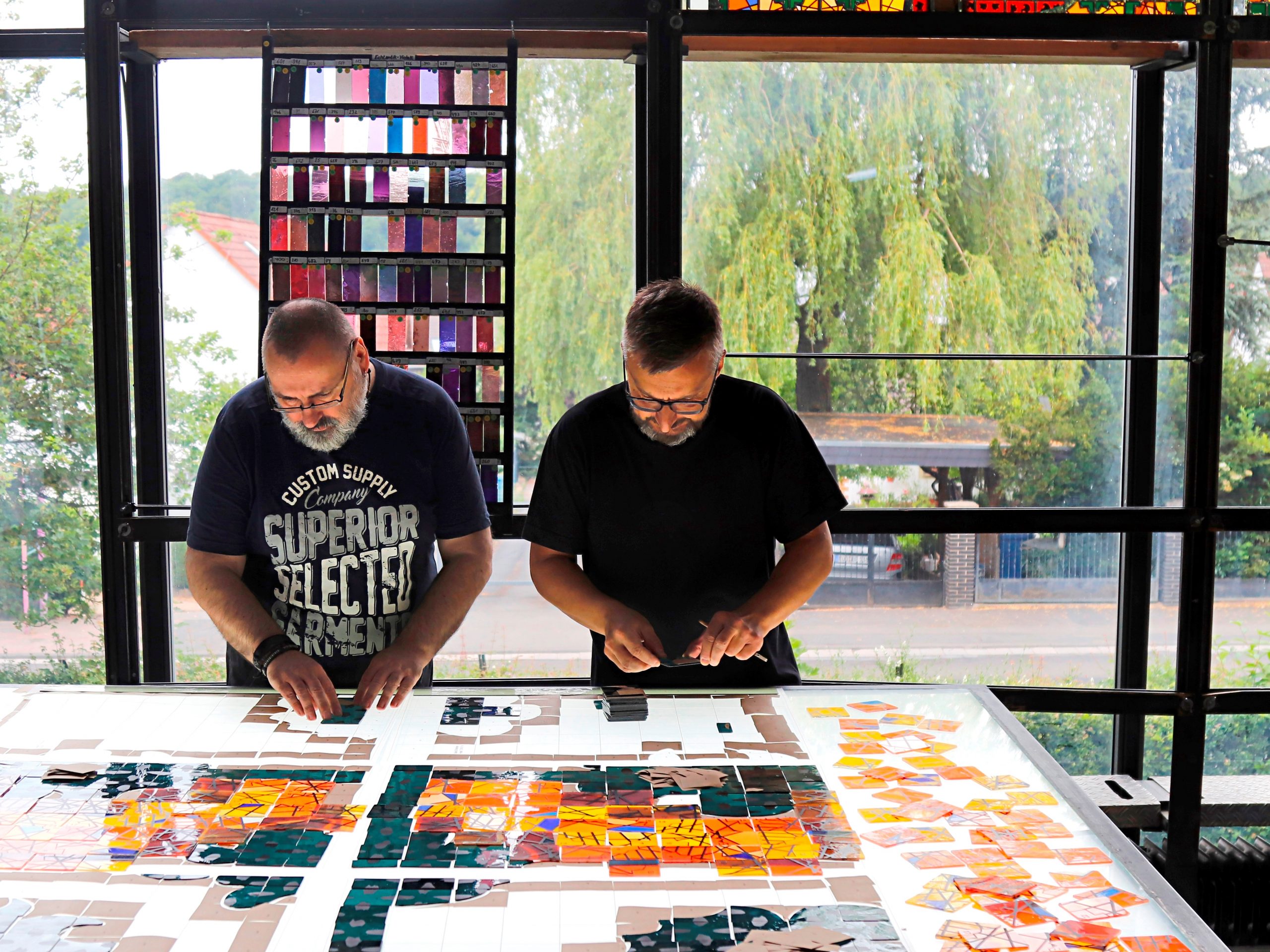 Zwei Personen arbeiten konzentriert an einem großen, farbigen Glasfenster in einem lichtdurchfluteten Atelier.