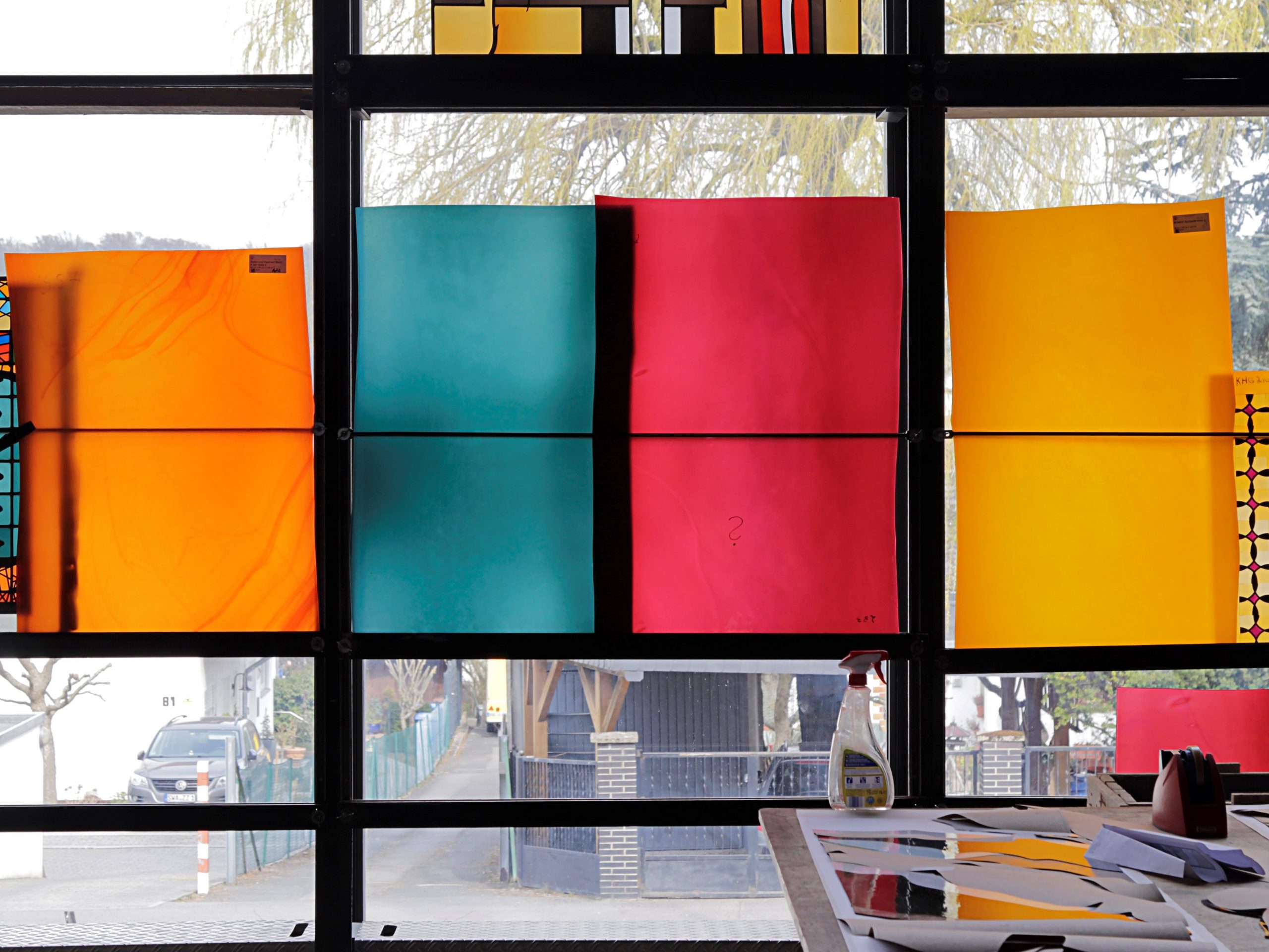 Große farbige Glasplatten in Orange, Türkis, Rot und Gelb vor einem Fenster in einer Glaswerkstatt.