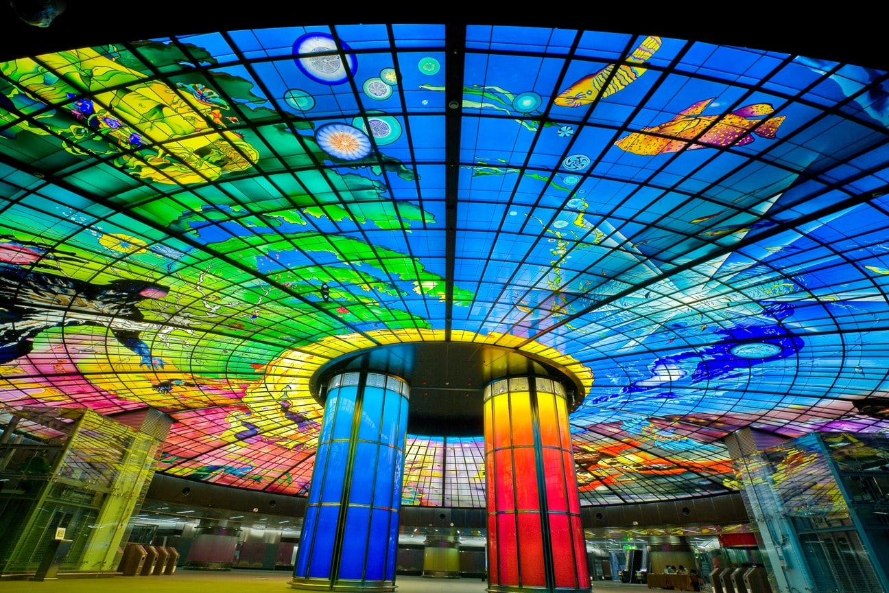 Farbenfrohes Glasdach mit abstrakten Motiven und kreisförmiger Struktur in einem modernen Gebäude in einer Bahnstation.