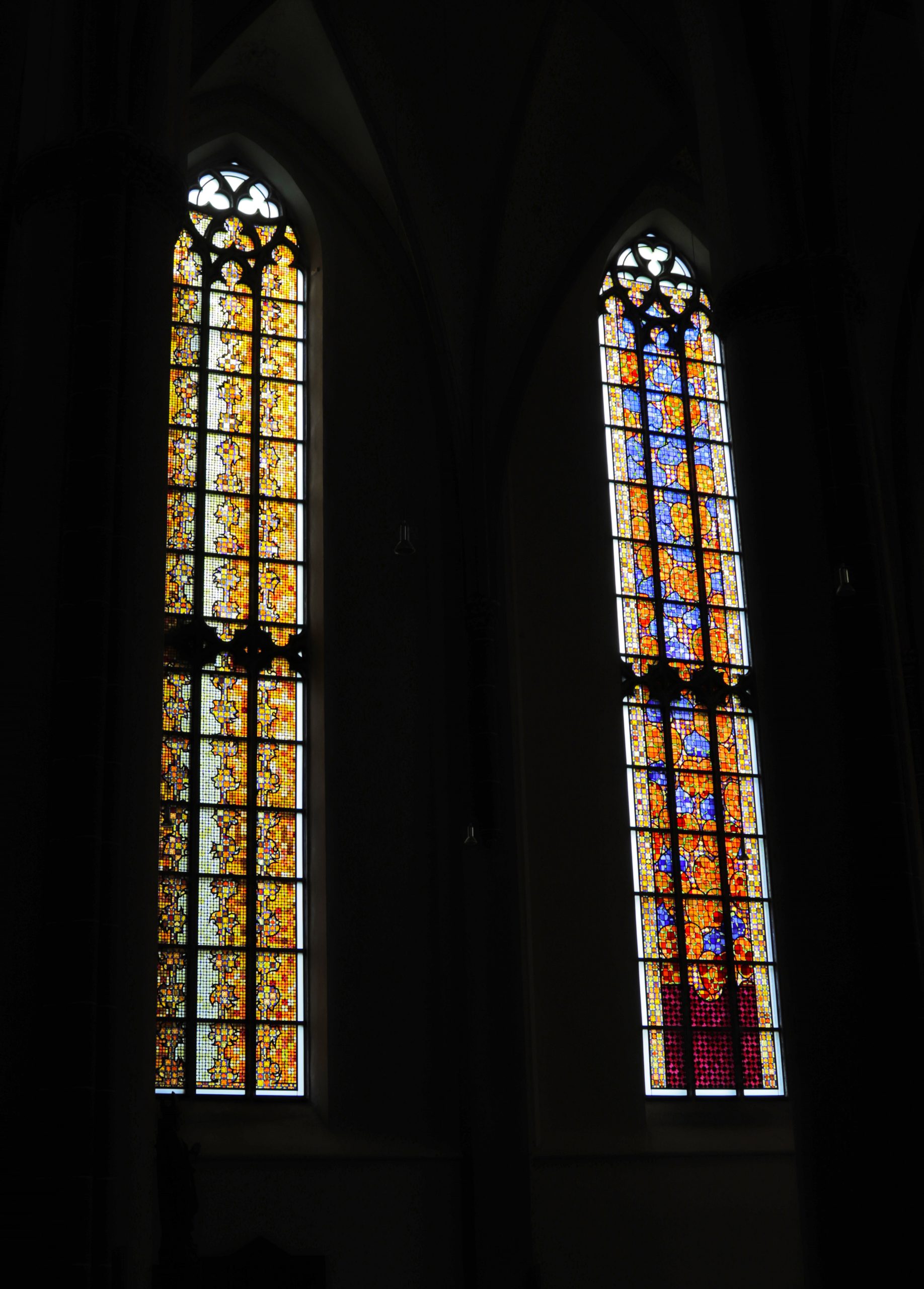 Zwei hohe gotische Kirchenfenster, gefüllt mit buntem Buntglas, das kunstvolle Muster und Motive zeigt, beleuchtet in einem dunklen kirchlichen Interieur.