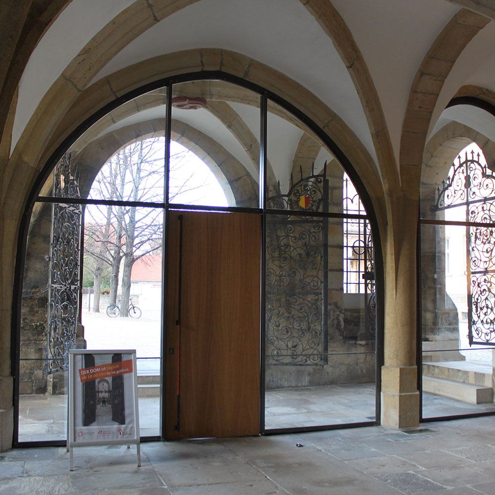 Blick durch eine geöffnete Holztür mit Glaspartien auf das Äußere eines historischen Gebäudes, eingefasst von steinernen Bögen.