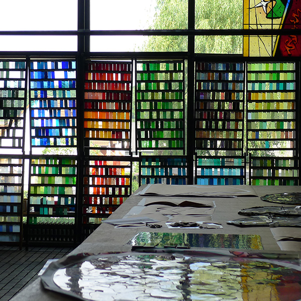 Blick in ein Atelier mit bunten Glasfenstern und diversen Glasstücken auf einem Tisch im Vordergrund.