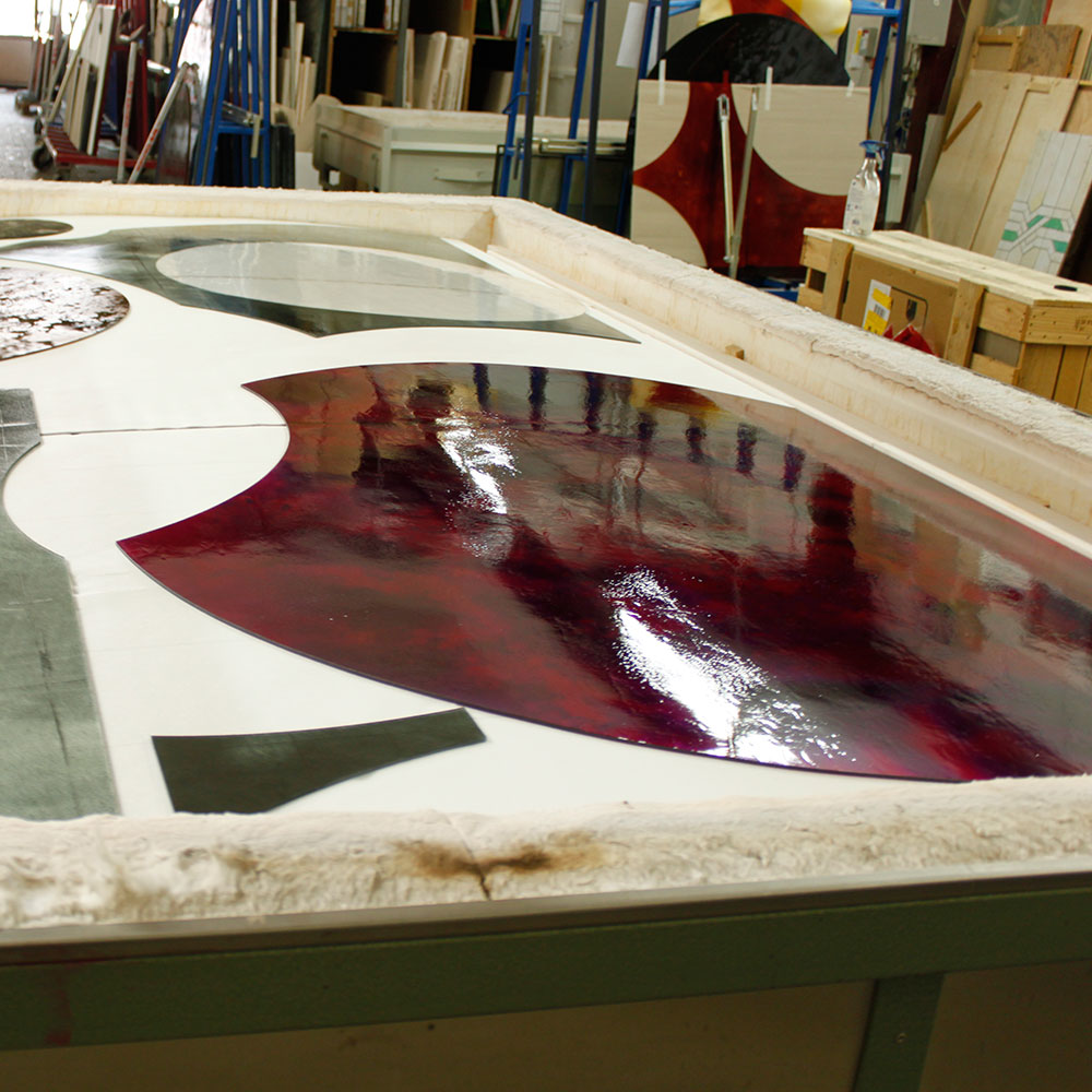 Ein großes, rotes, geschwungenes Glaselement wird in der Werkstatt bearbeitet.