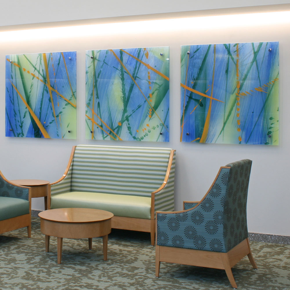 Drei abstrakte Glasgemälde mit blau-grünen und orangen Motiven über einer Sitzgruppe mit gestreiftem Sofa und Sesseln.