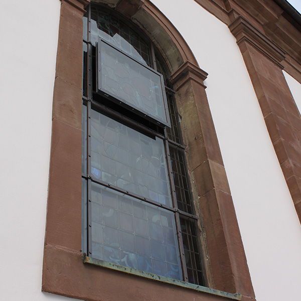 schutzverglasung_02 Schutzverglasung vor einem historischen Kirchenfenster, eingefasst in eine Steinmauer, bietet Schutz und Erhaltung für das dahinterliegende Kunstwerk.