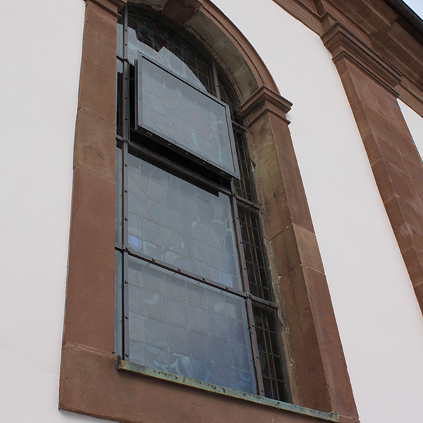 Schutzverglasung vor einem historischen Kirchenfenster, eingefasst in eine Steinmauer, bietet Schutz und Erhaltung für das dahinterliegende Kunstwerk.