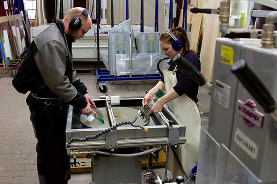 Zwei Personen arbeiten konzentriert an einer Glasbearbeitungsmaschine in einer Werkstatt. Sie Schleifen scheinbar Kanten von Scheiben.