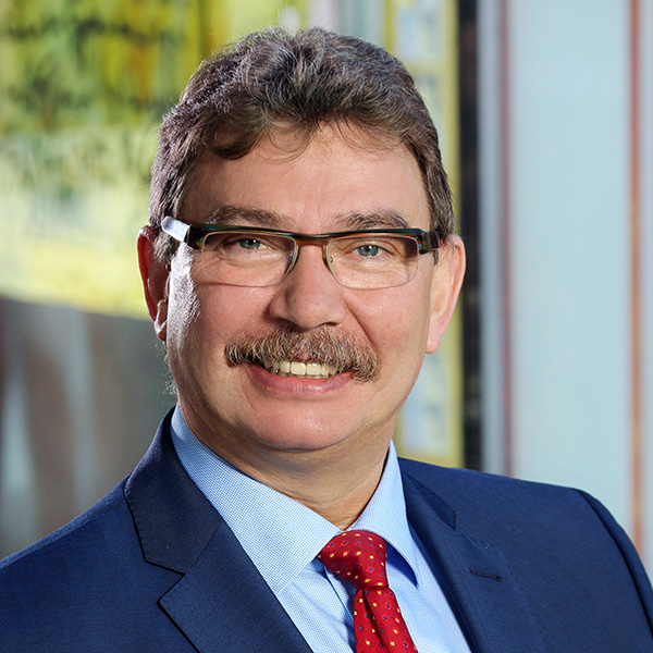 Rainer Schmitt: Portrait of a smiling man with a moustache, wearing glasses, a blue jacket and a red tie against a blurred background.