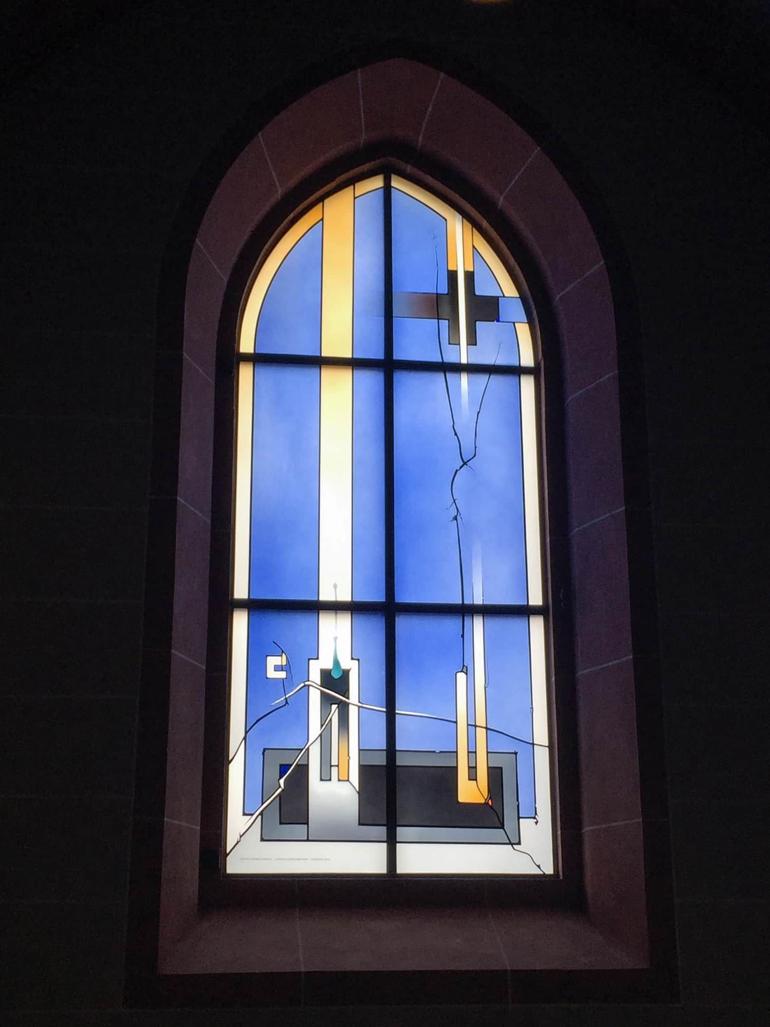 Farbenreiches Kirchenfenster mit modernem Design in Blau- und Gelbtönen innerhalb gotischer Bogenstruktur.