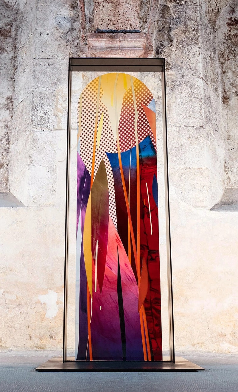 Moderne Glaskunstinstallation mit lebhaften Farben und geometrischen Mustern in einem Metallrahmen, ausgestellt vor einer Steinwand.