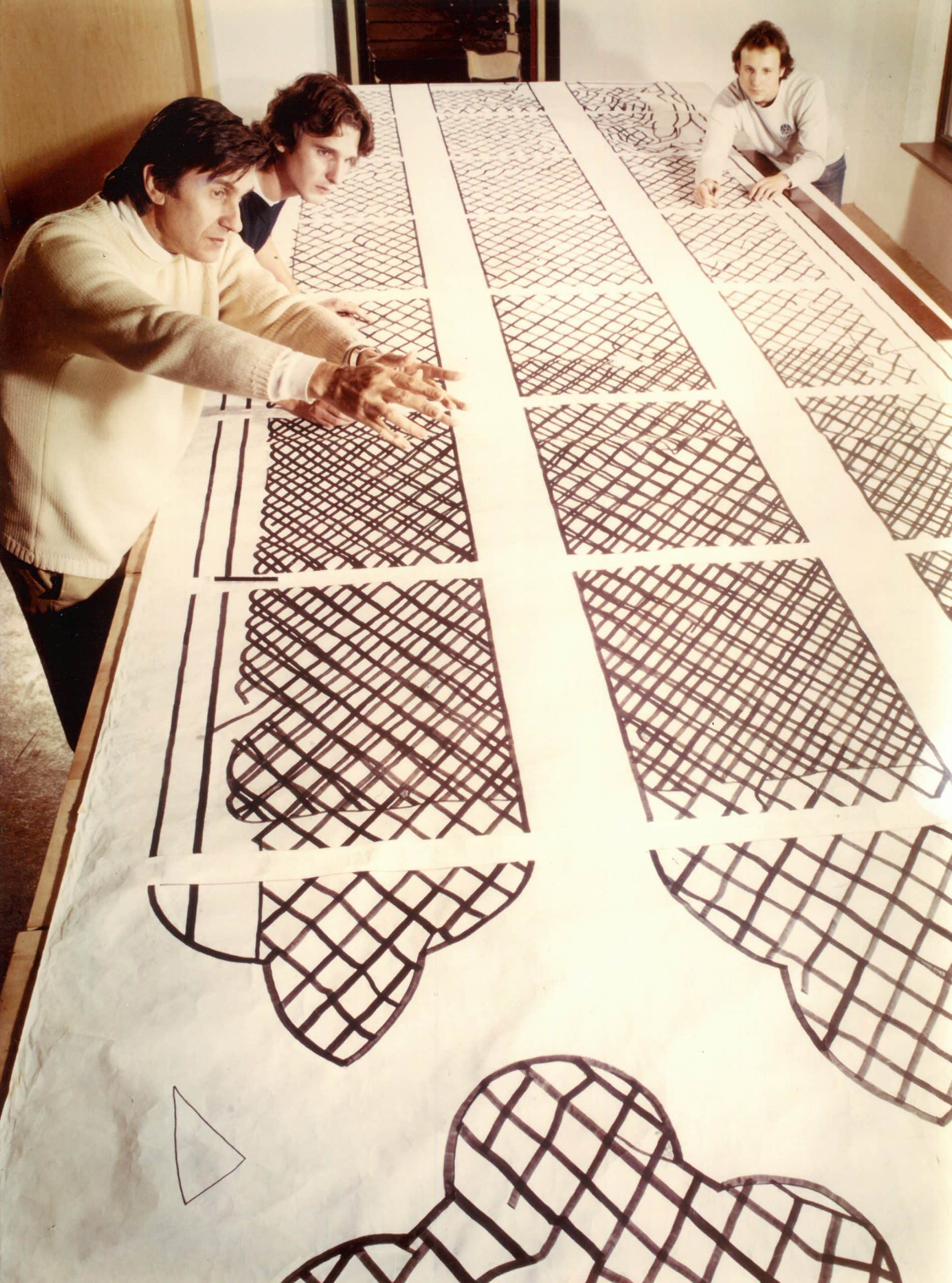 Vergilbtes Foto: Drei Personen betrachten Entwurfszeichnungen für Glaskunstfenster in einem Atelier.