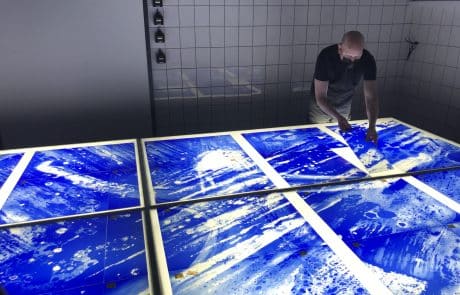 Person überprüft mehrere große blaue Glasplatten mit abstrakten Mustern auf einem Tisch in einem Atelier.