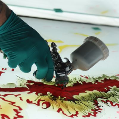 Hand in türkisen Handschuhen benutzt Airbrush auf einem bunten Glaskunstwerk, Kunsthandwerk, detailreiche Farbgebung.