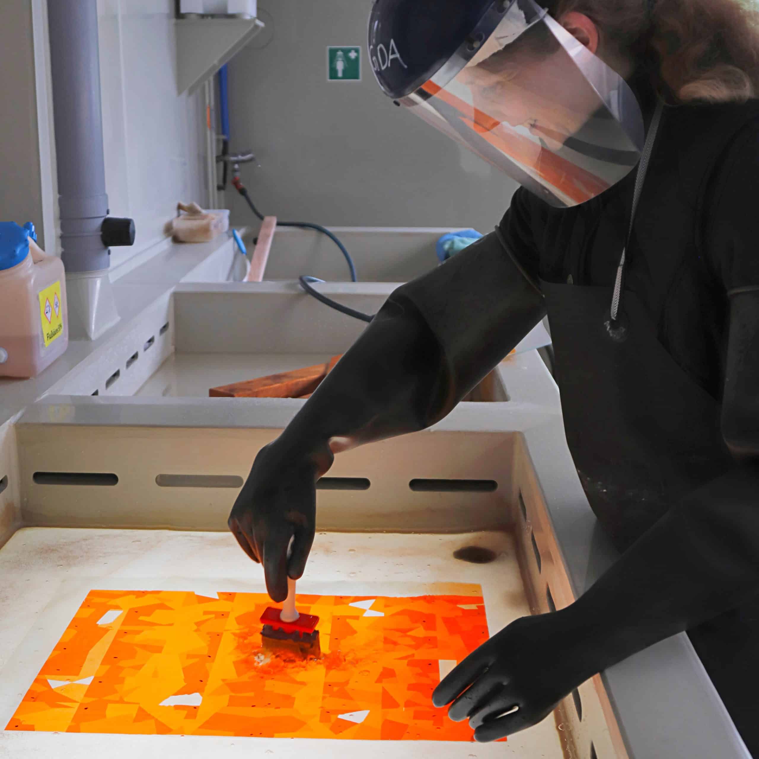 Person in Schutzkleidung arbeitet an einem leuchtend orangefarbenen Glasmosaik in einem Atelier.