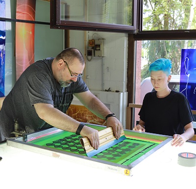 Zwei Personen arbeiten konzentriert in einem Atelier an einem farbigen Glasfenster, mit Werkzeugen (Rakel) auf einem beleuchteten Tisch.