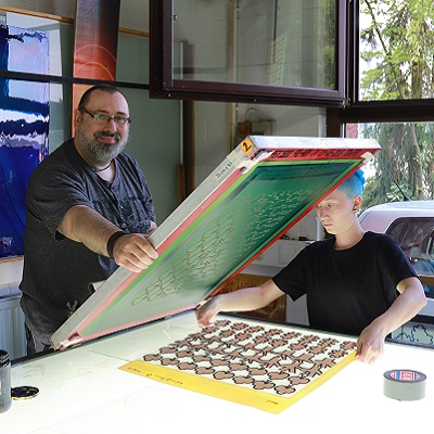 Zwei Personen arbeiten sorgfältig an einem farbigen Glasobjekt in einem Künstleratelier. Es zeigt das Siebdruckverfahren für Glasscheiben.