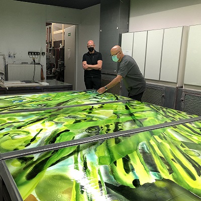 Two people look at a large, colorful glass work of art with green and yellow patterns on a work table.