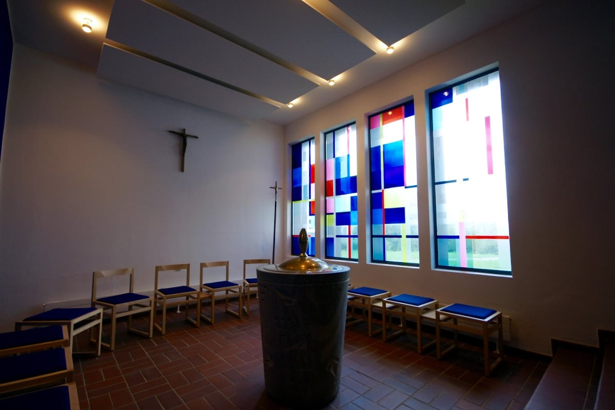 Modern gestaltetes Kircheninterieur mit bunten Glasfenstern, Taufbecken im Vordergrund, Kreuz an der Wand und Holzbänken.