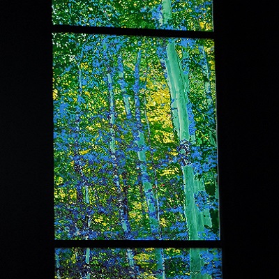 Colorful glass windows with forest motif in blue and green tones, artwork illuminated against dark background.