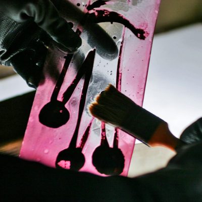 A craftsman carefully removes glass overtaking with a brush on an illuminated, colored glass piece. Glass etching.