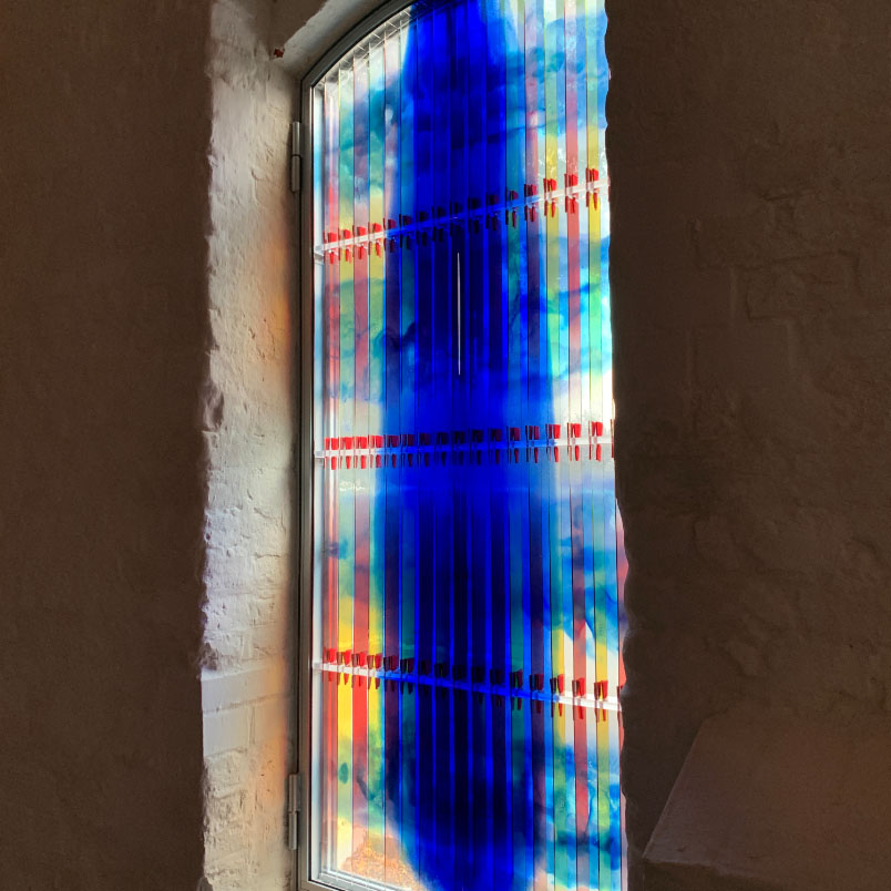 Farbenfrohes Kirchenfenster mit blauen und roten Akzenten, das Sonnenlicht durchlässt und leuchtende Reflexionen an der Wand erzeugt.