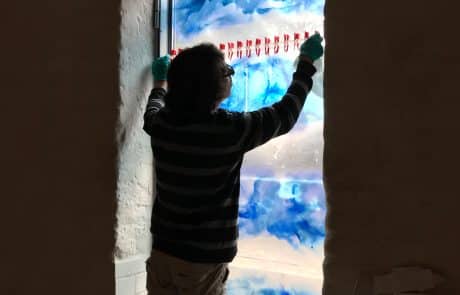 Person befestigt ein farbenfrohes Glasfenster mit blauen und roten Elementen an einem Wanddurchbruch.