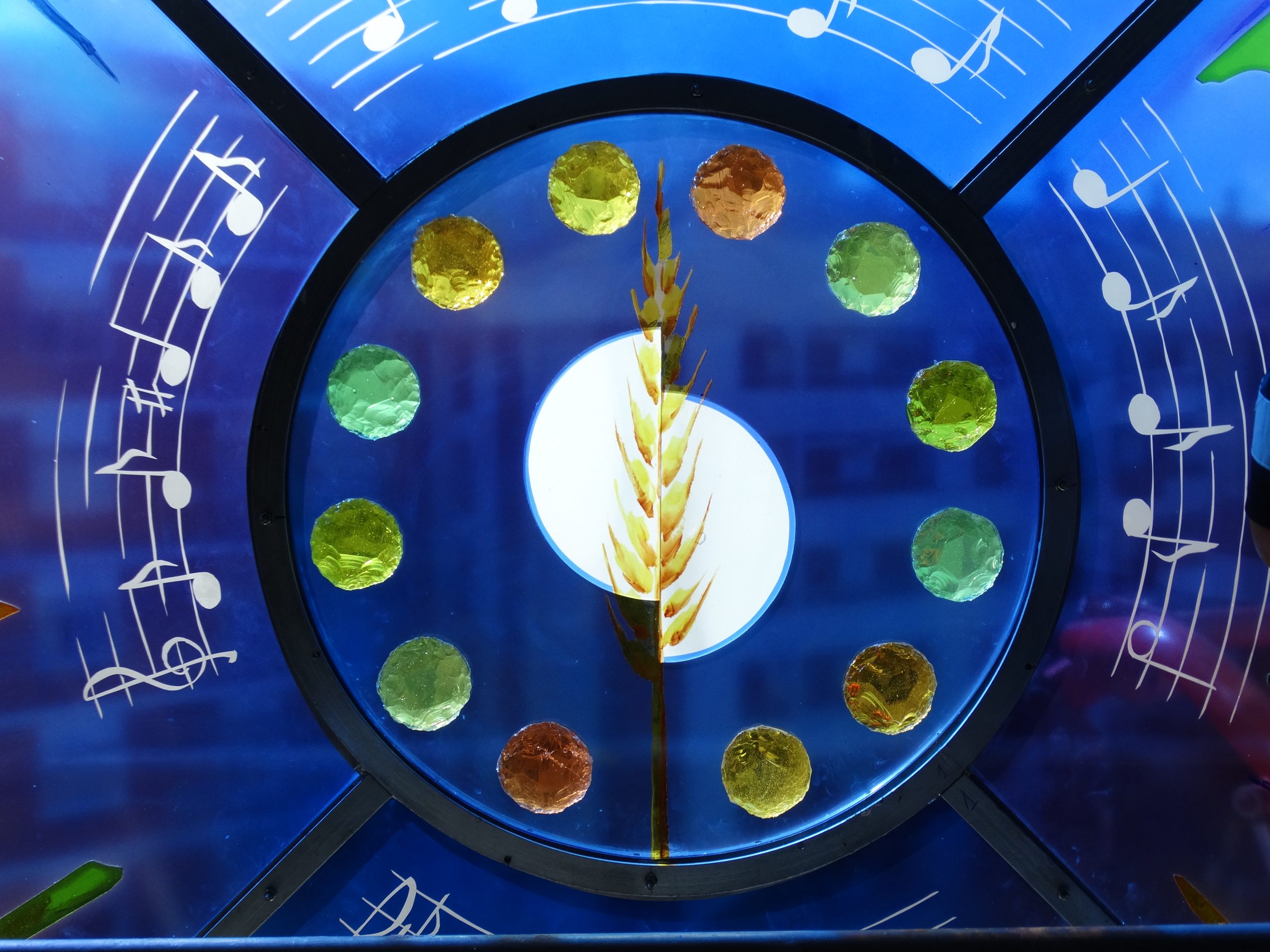 Kunstvolles Buntglasfenster mit blauem Hintergrund, eingelassenen runden Glaselementen und Weizenähre im Zentrum und Musiknoten.