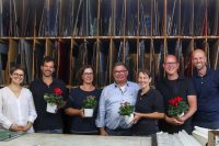Sechs lächelnde Personen in einem Atelier, halten Blumen und stehen vor Regalen mit bunten Glasplatten.