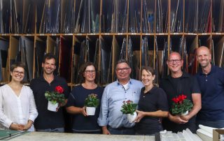 Sechs lächelnde Personen in einem Atelier, halten Blumen und stehen vor Regalen mit bunten Glasplatten.