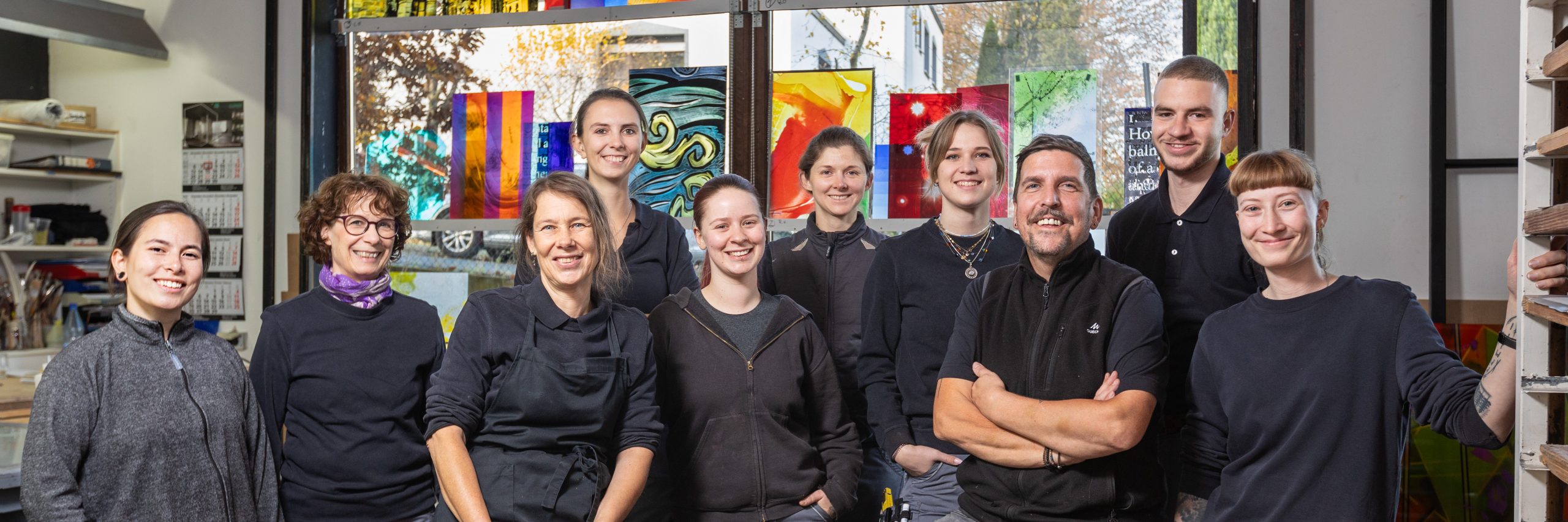 Gruppe lächelnder Mitarbeiter vor bunten Glaskunstwerken im Derix Glasstudio, welches die Vielfalt und Kreativität in der Gestaltung mit Glas zeigt.