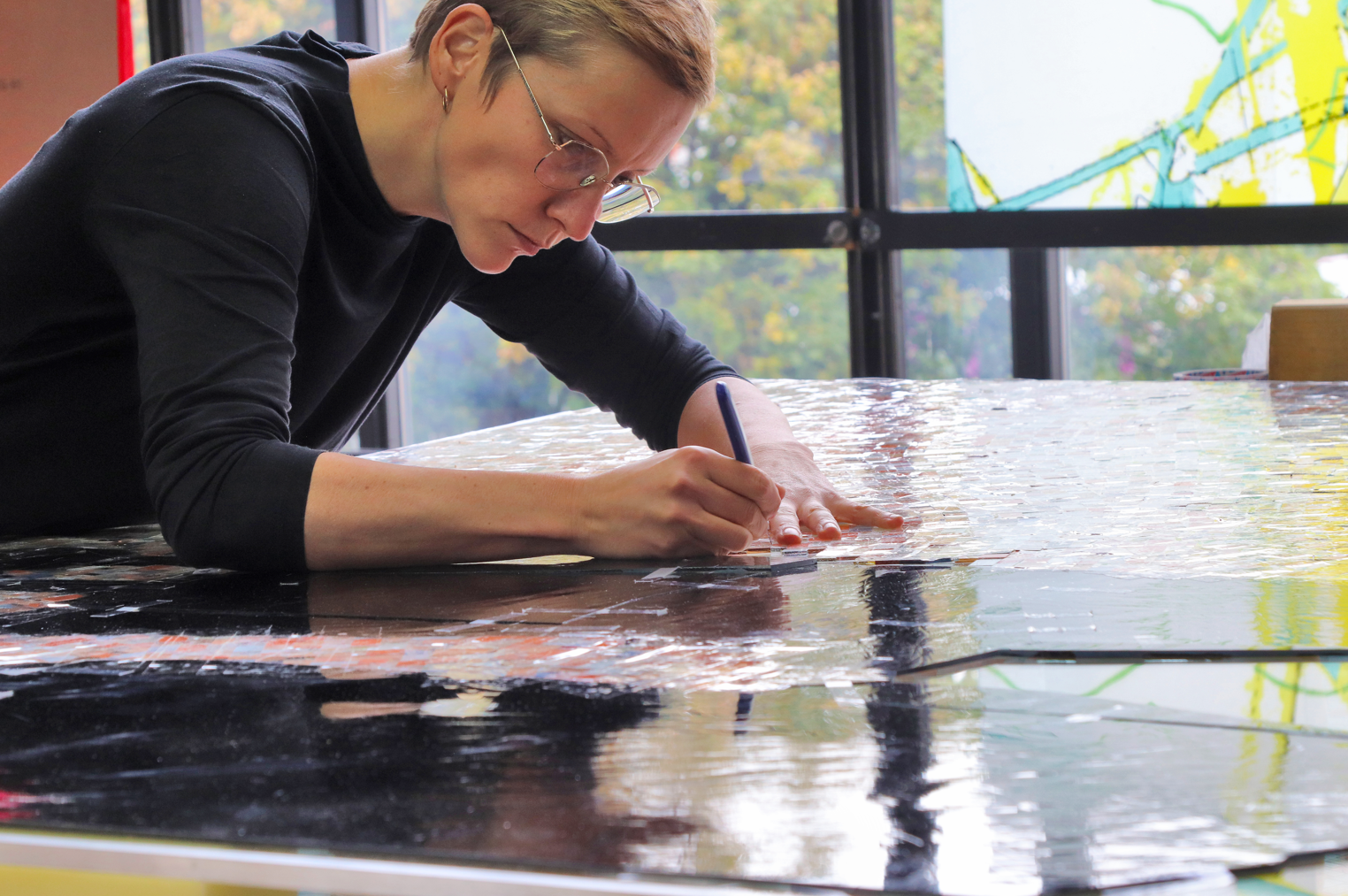 Glaskünstlerin arbeitet konzentriert an einem großen, farbigen Glasfenster in einem hell erleuchteten Atelier.