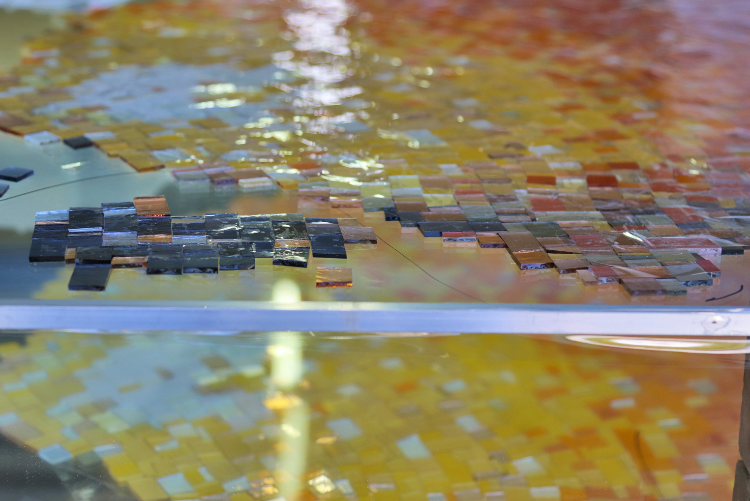 Detailaufnahme eines farbenfrohen Glaskunstwerks, gezeigt in Form eines Mosaiks aus zahlreichen kleinen, quadratischen Glasstücken in verschiedenen Schattierungen von Gelb, Orange und Schwarz.