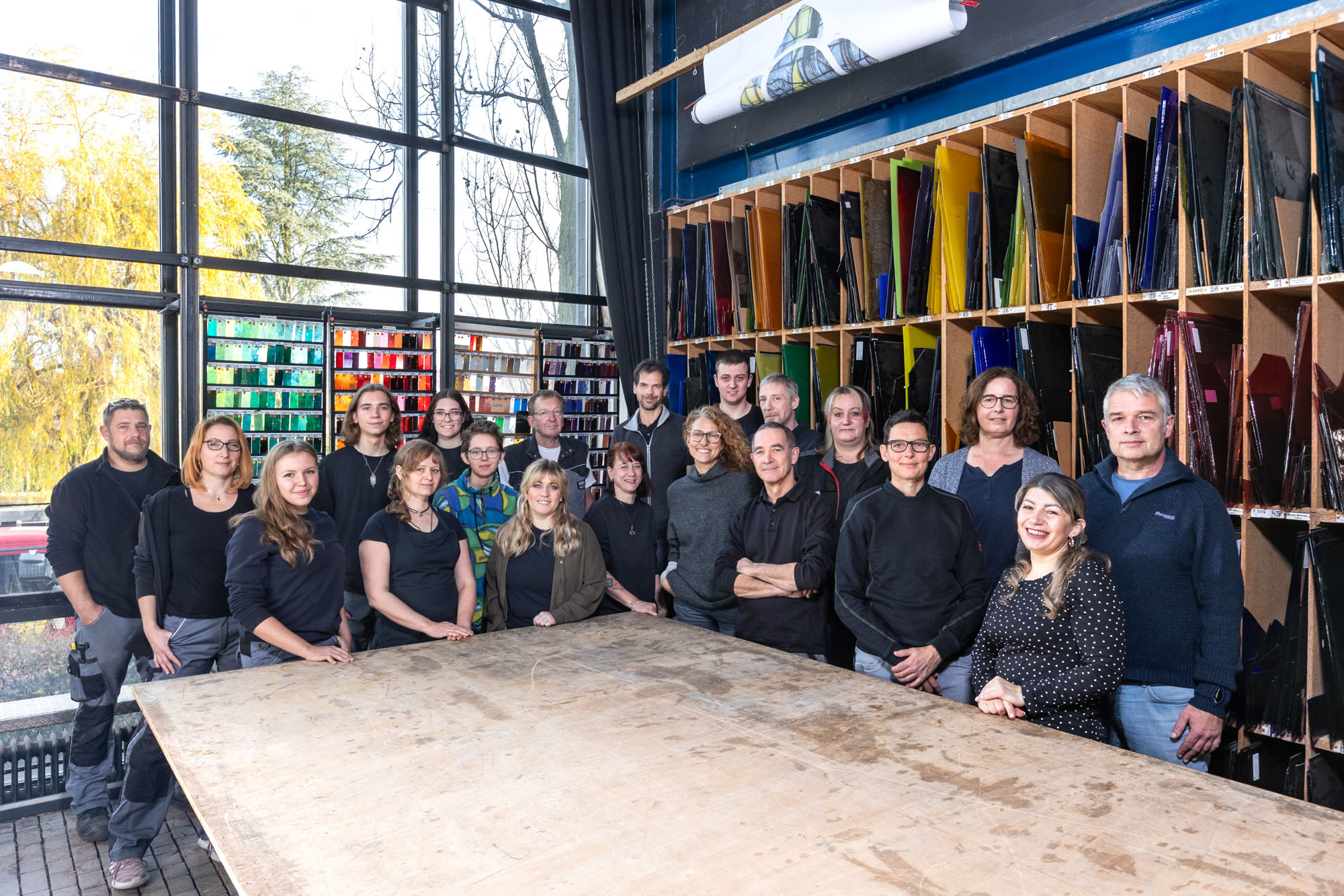Mitarbeiter Glaserei copy Team von Glas-Künstlern in einer Werkstatt mit farbenfrohen Glasplatten im Hintergrund.