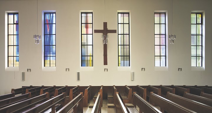 Derix_Fenster_rechts_5-1-bearb – Derix Glasstudios Innenansicht einer Kirche mit modernen Buntglasfenstern, die abstrakte, farbige Muster zeigen, und einem zentralen Kreuz über einem Altar, umgeben von Holzbänken.