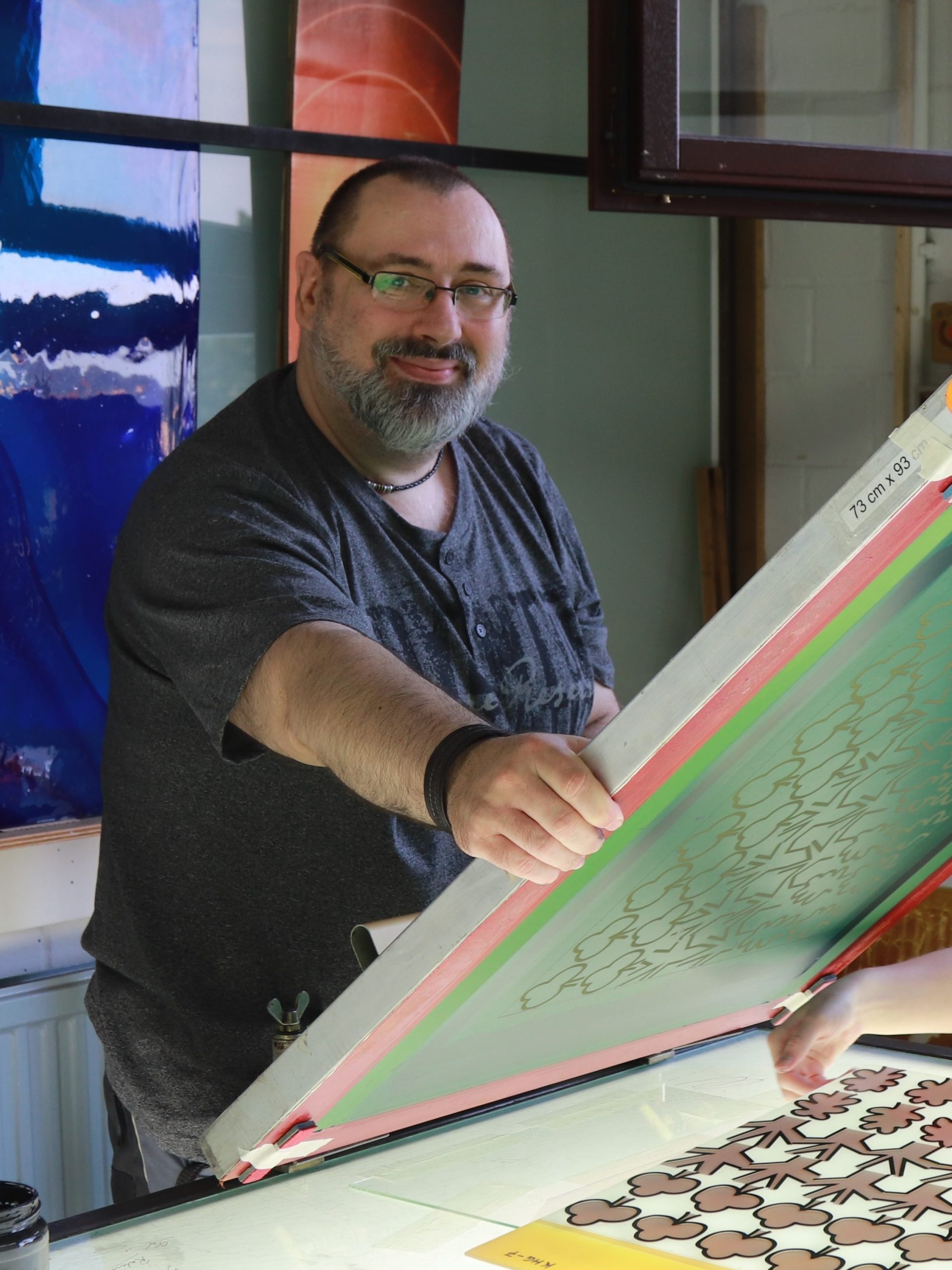 Ein Mann in einem Atelier hält eine Siebdruckvorlage vor farbenfrohen Glaspaneelen.